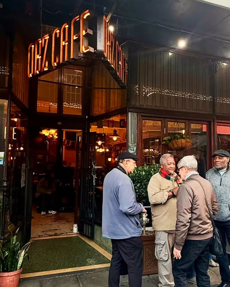 Regulars chatting outside Obz Café under the copper sign on the corner of Lower Main Road